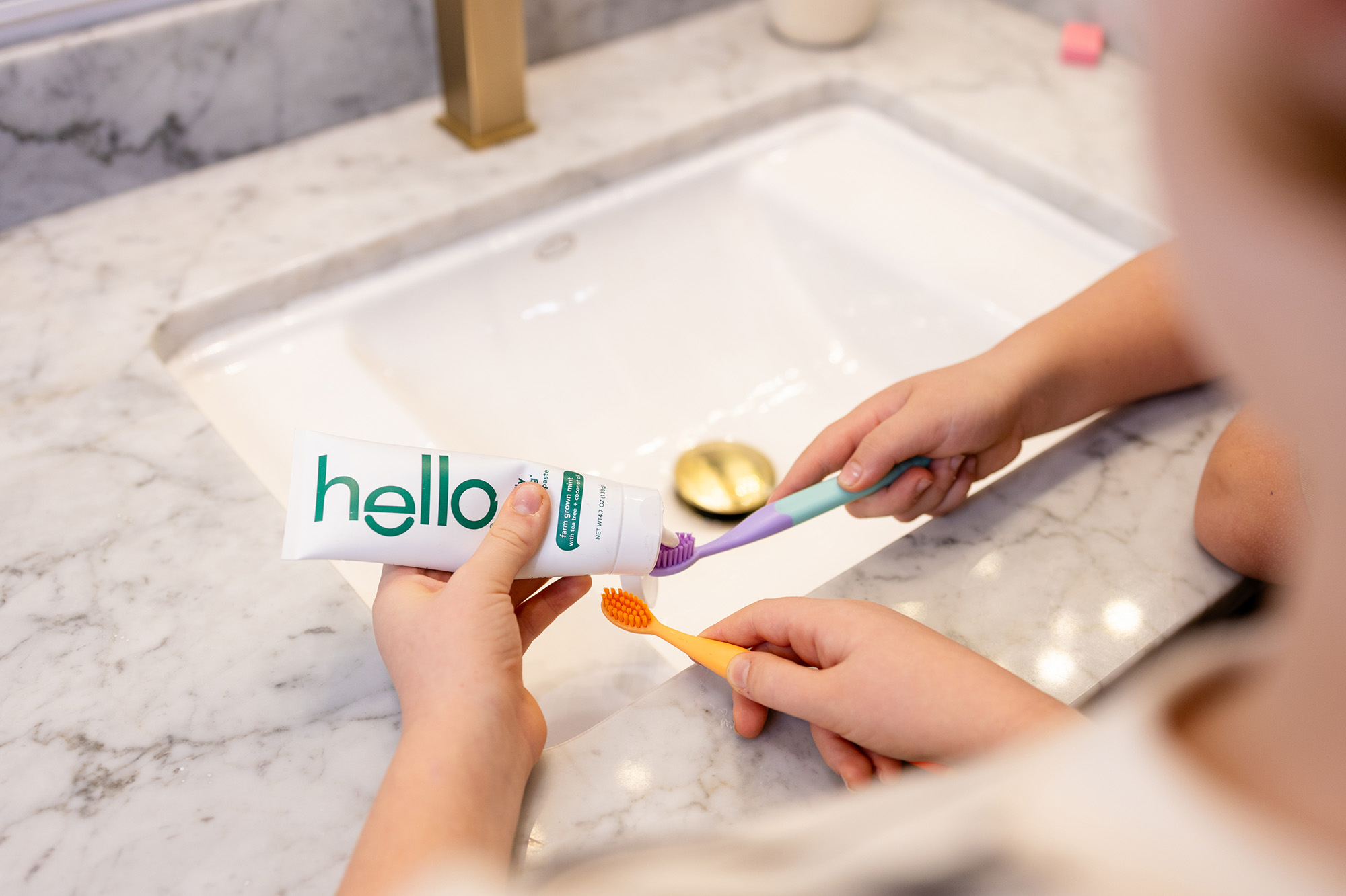 Two children holding toothbrushes and squeezing toothpaste from a Hello brand tube over a bathroom sink.